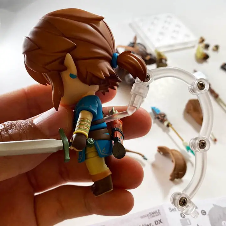 A hand holding a Link figure with a plastic peice coming out of its back with a bunch of parts in teh backgorund (link nendodorid unboxing)
