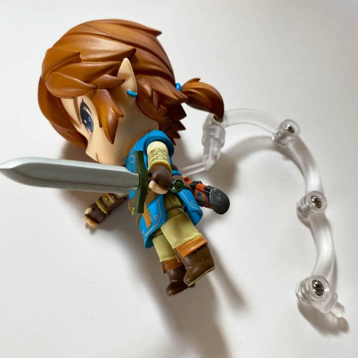 Link lying on a white tabletop with a peice of clear plastic jutting from his back (link nendodroid unboxing)