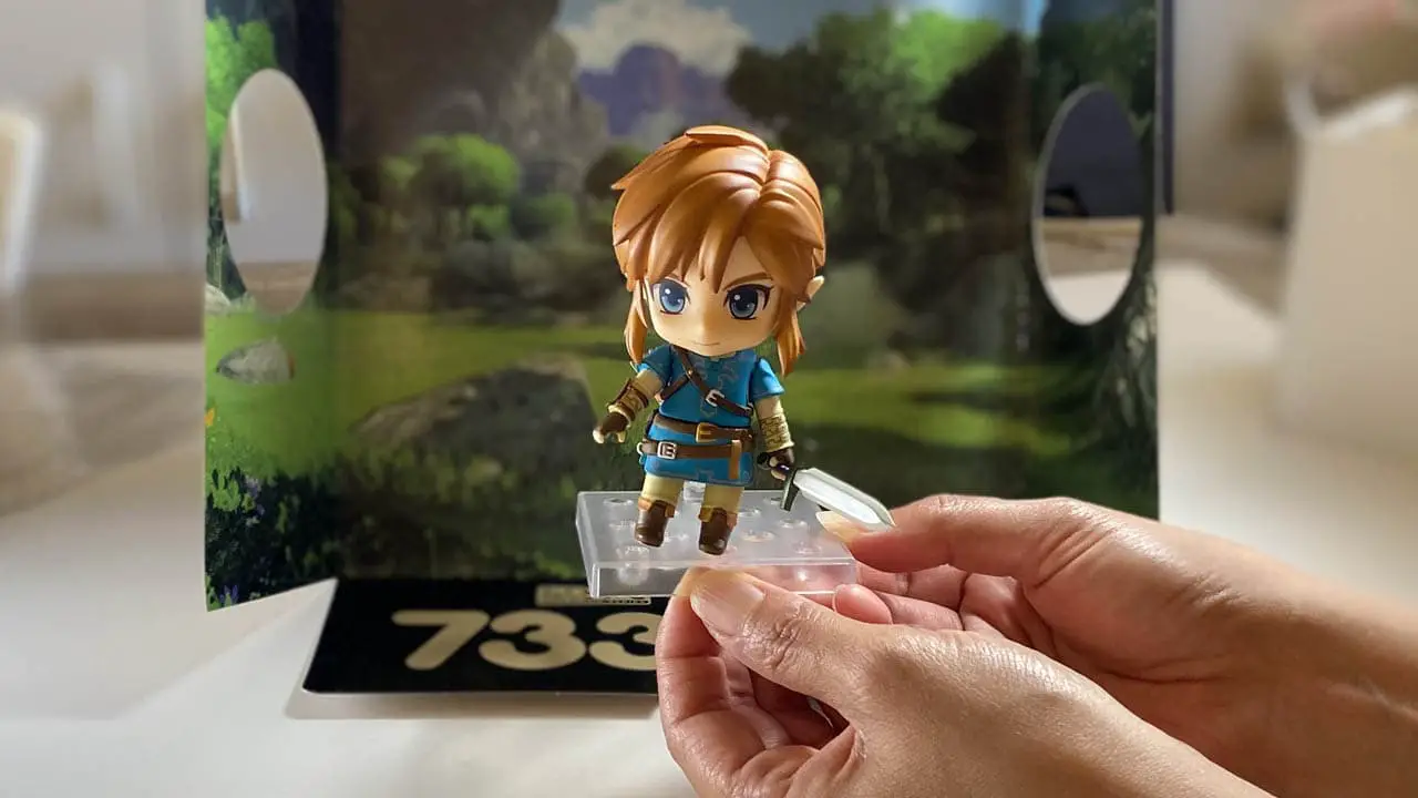 Hands holding link figure in front of a forest display atop a table (link nendoroid unboxing)